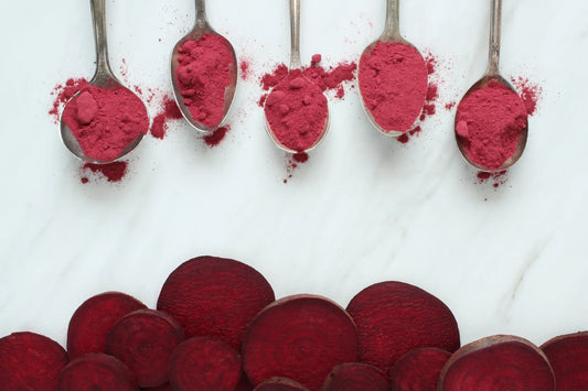 Multiple spoons filled with beetroot powder and slices of natural beets arranged on a granite countertop.